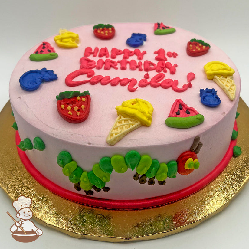 Round cake with pink icing, decorated with colorful fruits, ice cream cones, and a caterpillar design around the side.