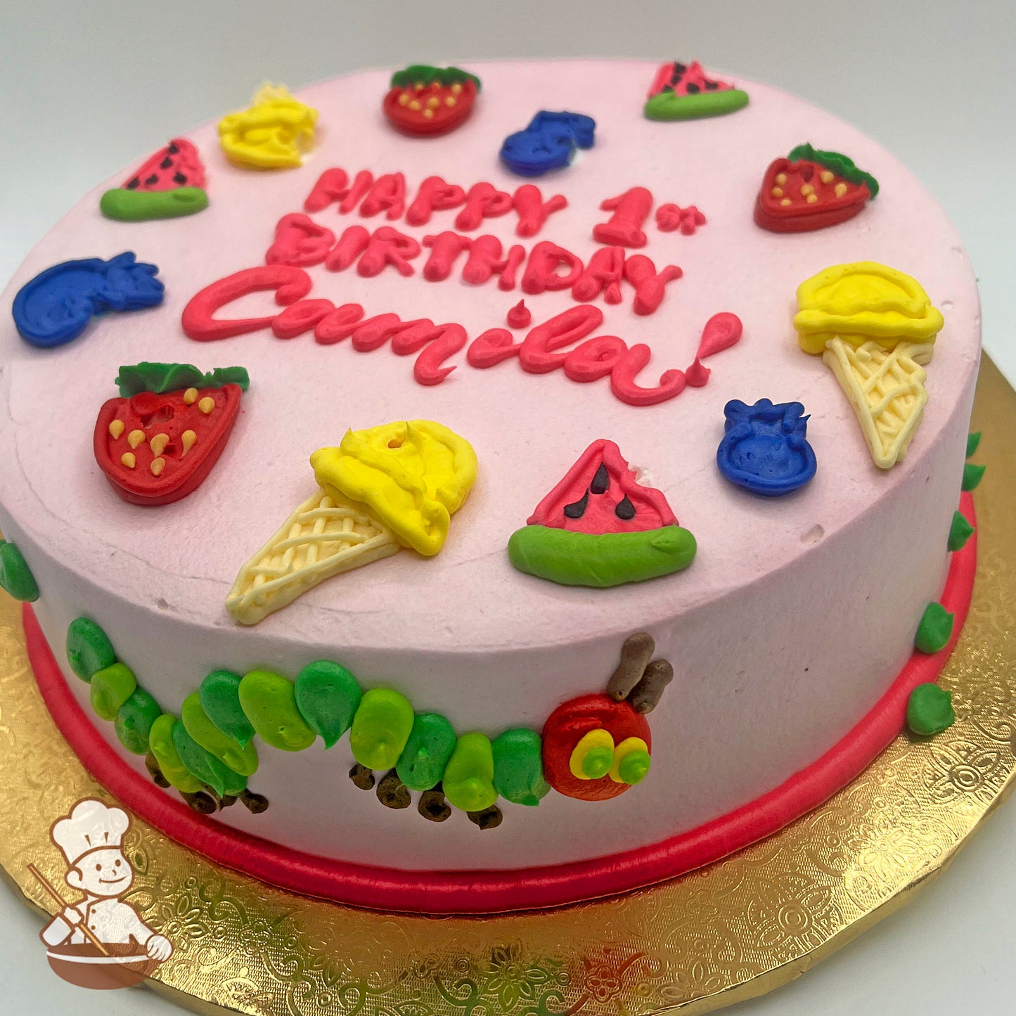 Round cake with pink icing, decorated with colorful fruits, ice cream cones, and a caterpillar design around the side.