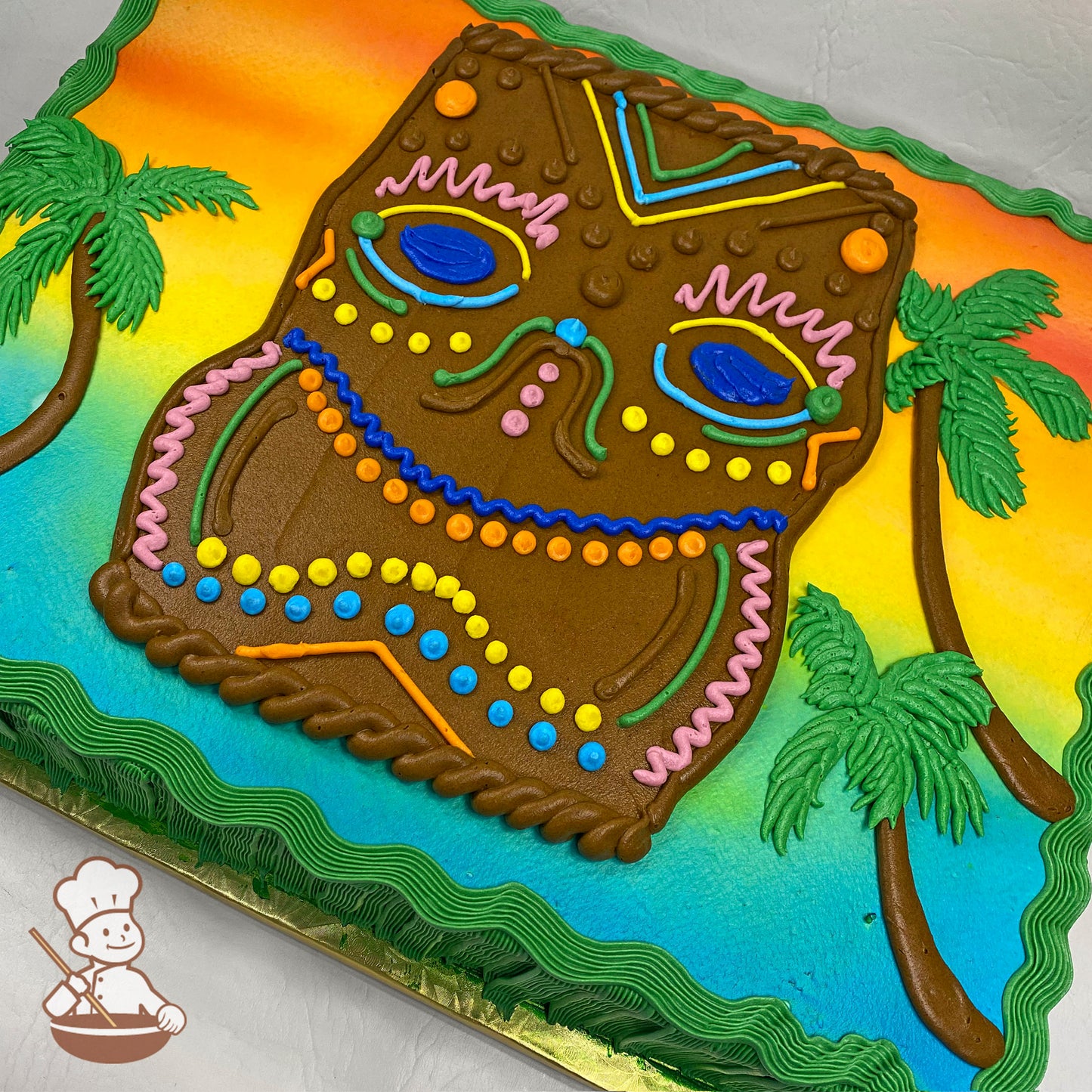 Sheet cake with a colorful sunset background, a large brown Tiki mask design decorated with vibrant icing details, and green palm trees.