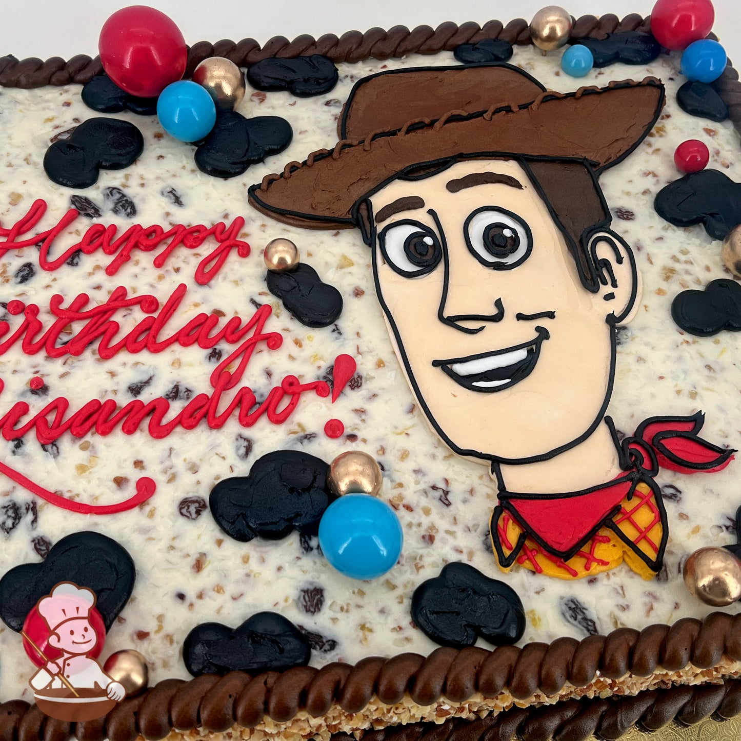 Sheet cake with a Woody-themed design, featuring a cowboy hat, cow print buttercream, red and blue plastic balloons, and gold spherical decorations.