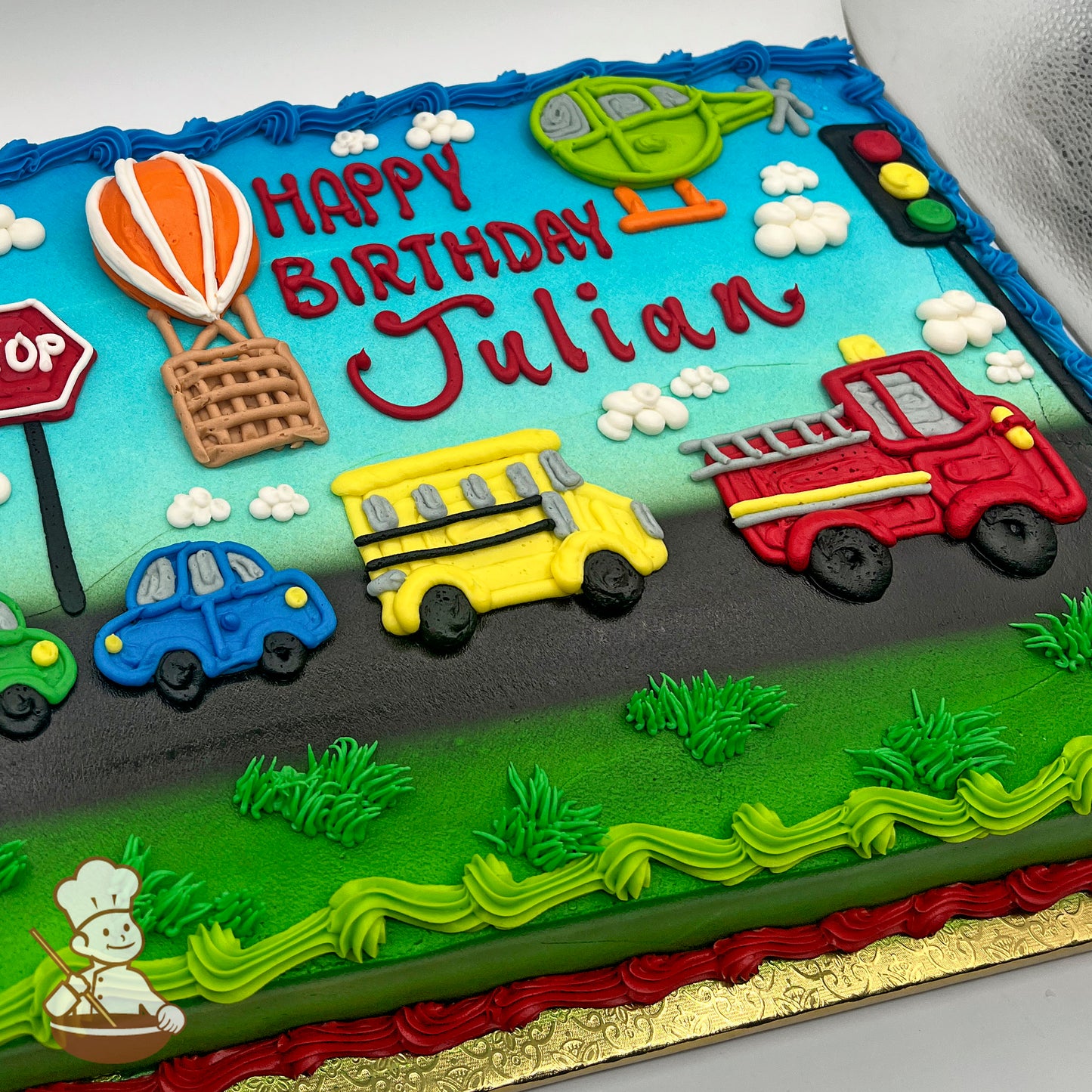 Sheet cake with buttercream decorations of a helicopter, hot air balloon, fire truck, school bus, cars, stop sign, stoplight, clouds, grass, and a road.