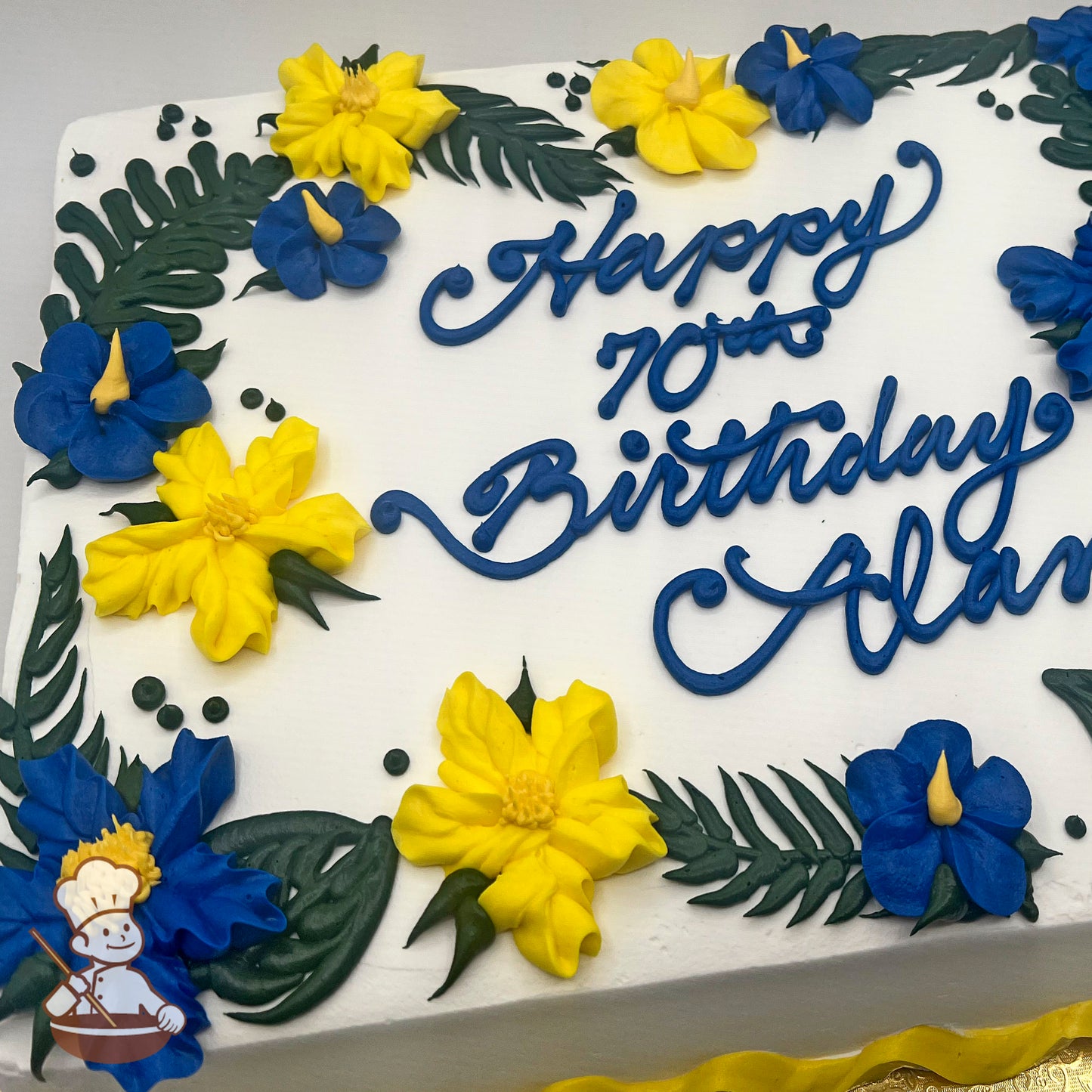 White sheet cake with buttercream decorations of yellow hibiscus flowers, blue tropical flowers, green fern leaves, and small green dots.