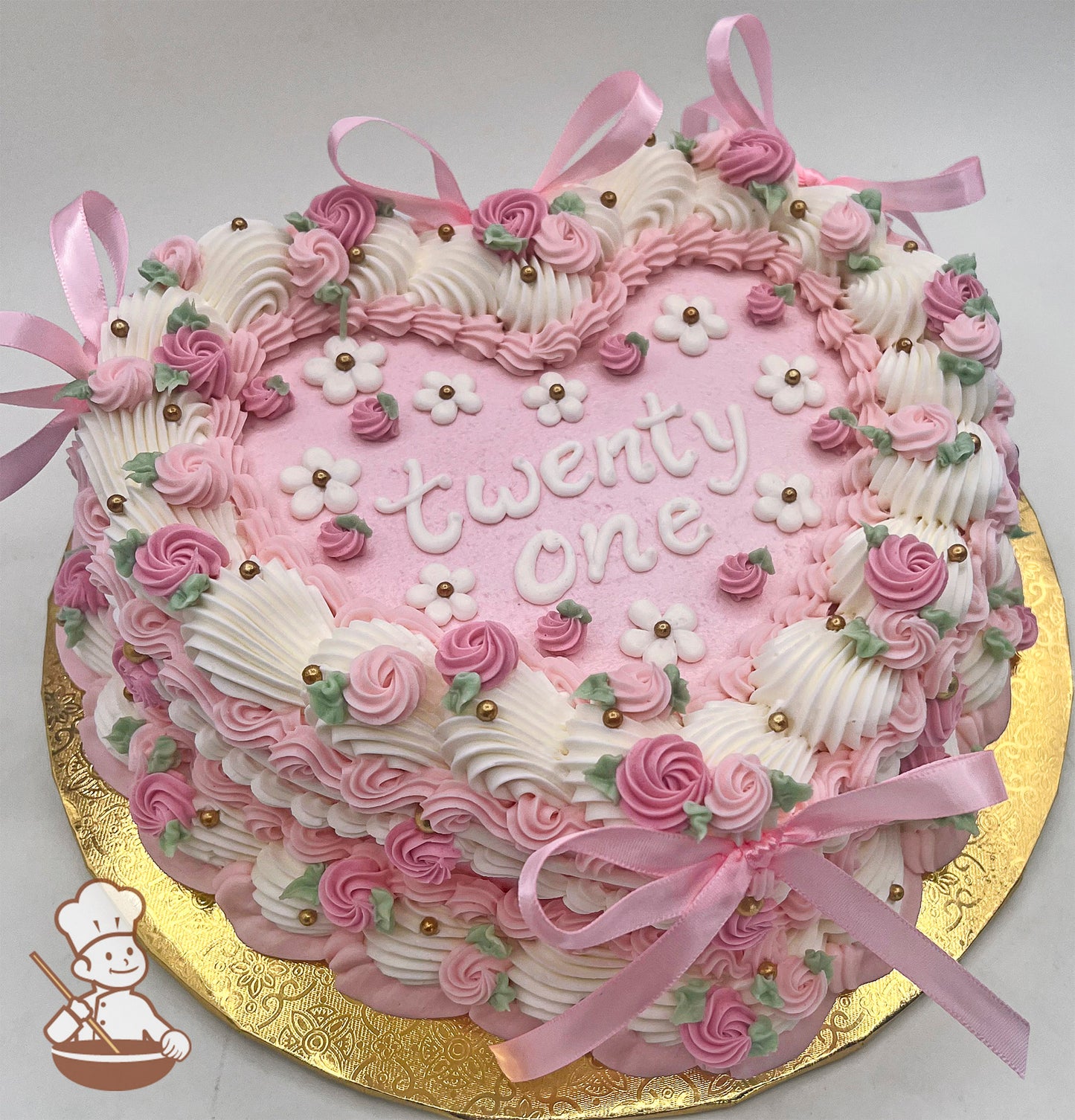 Heart-shaped pink sheet cake with buttercream rosettes, white flowers, gold sugar pearls, green leaves, and pink satin bows on a gold base.