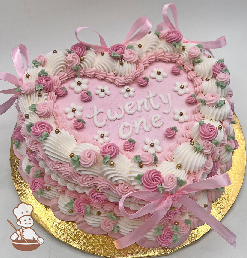 Heart-shaped pink sheet cake with buttercream rosettes, white flowers, gold sugar pearls, green leaves, and pink satin bows on a gold base.