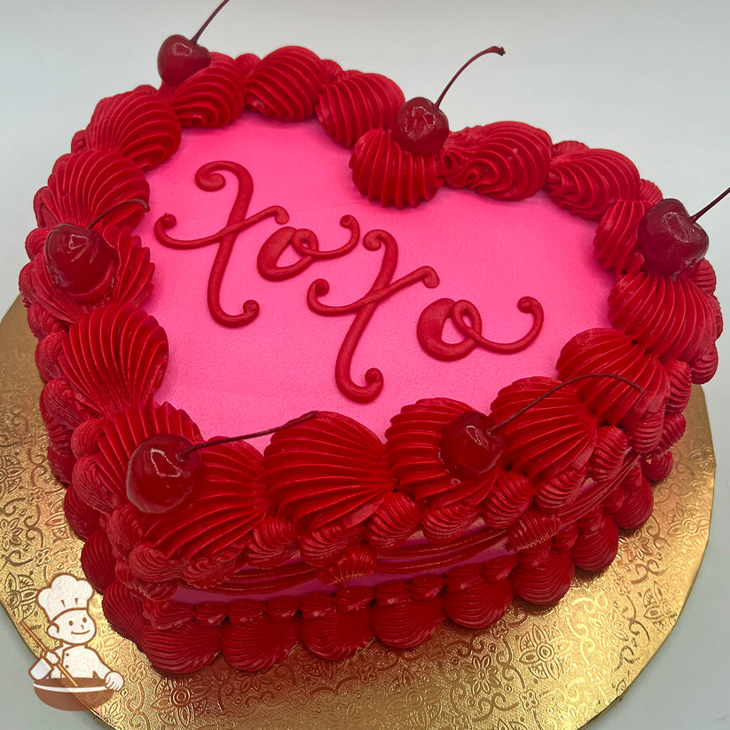 Heart-shaped cake with pink frosting, red buttercream piping, maraschino cherries on top, and a decorative gold base.
