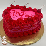 Heart-shaped cake with pink frosting, red buttercream piping, maraschino cherries on top, and a decorative gold base.