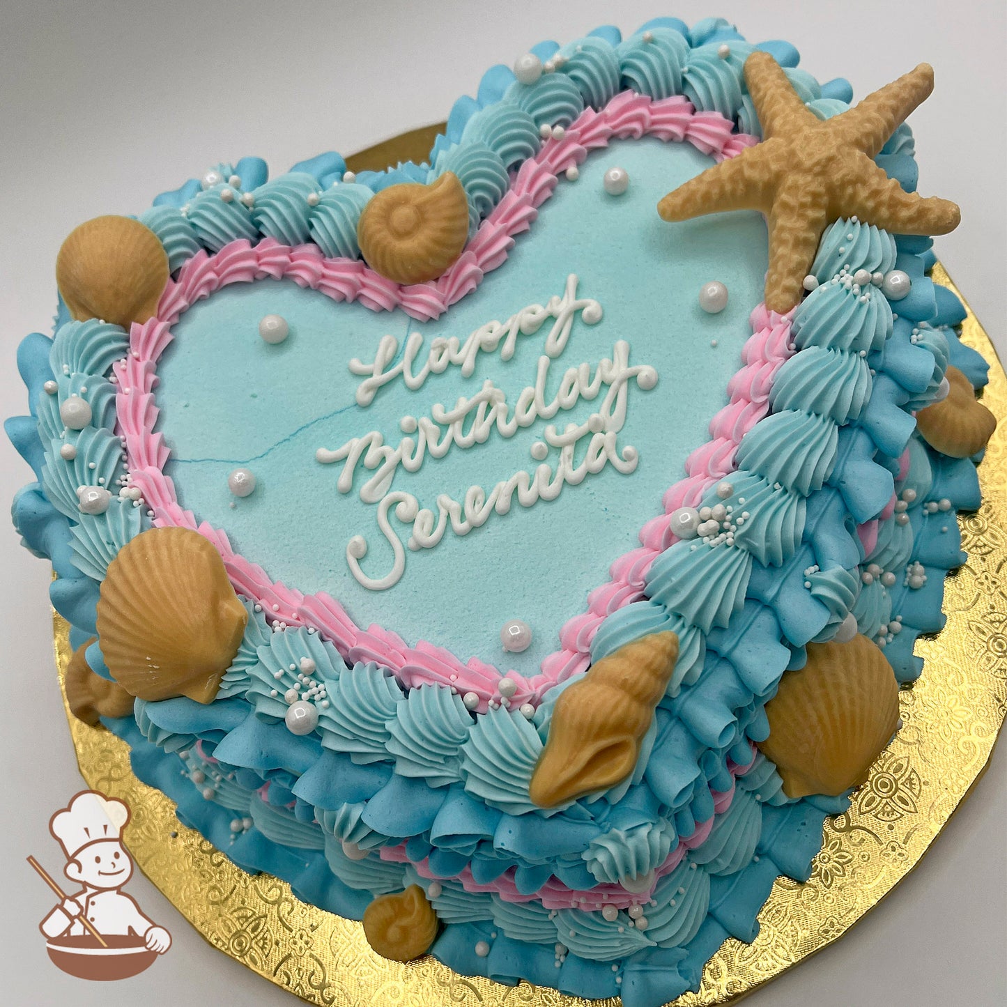 Heart-shaped cake with blue and pink vintage-style buttercream piping, white chocolate seashells and starfish and sugar pearls decoration on a gold board.