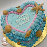 Heart-shaped cake with blue and pink vintage-style buttercream piping, white chocolate seashells and starfish and sugar pearls decoration on a gold board.