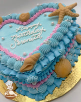 Heart-shaped cake with blue and pink vintage-style buttercream piping, white chocolate seashells and starfish and sugar pearls decoration on a gold board.