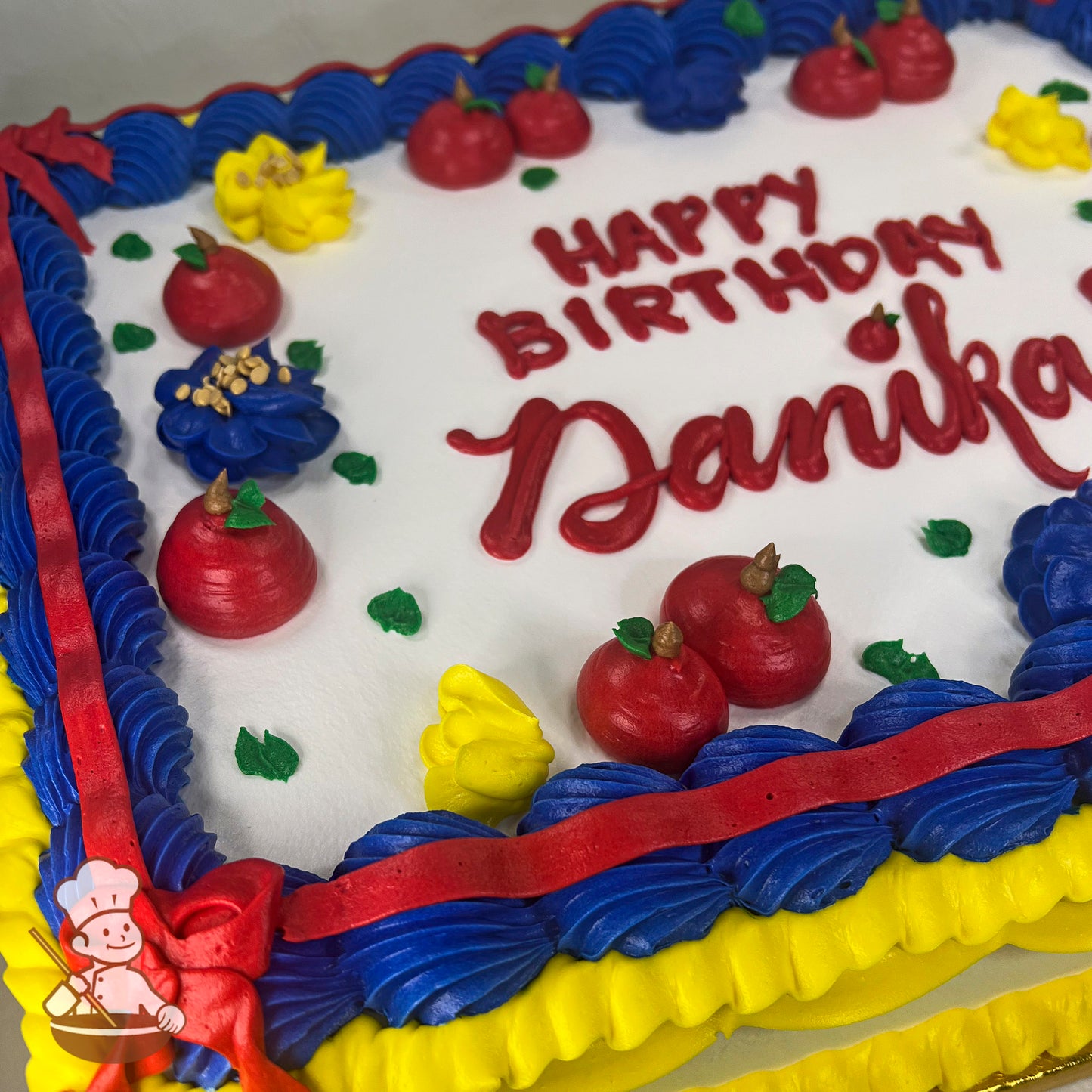 A sheet cake with vibrant buttercream decorations, featuring red apples, yellow flowers, blue flowers with yellow centers, green leaves, a red bow, and blue and yellow piped borders.
