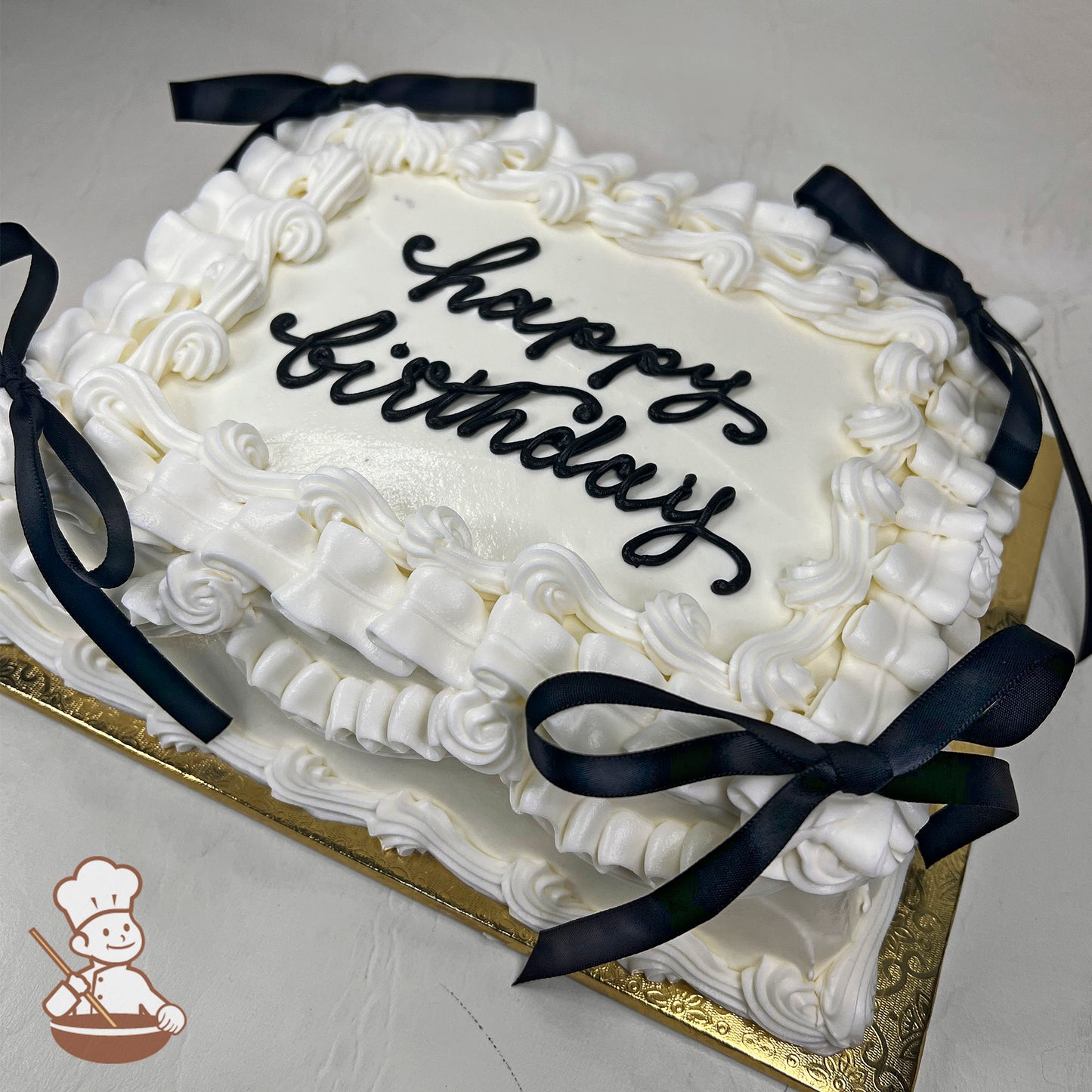 White sheet cake with intricate buttercream piping, ruffled edges, and black satin bows at the corners, placed on a gold tray.