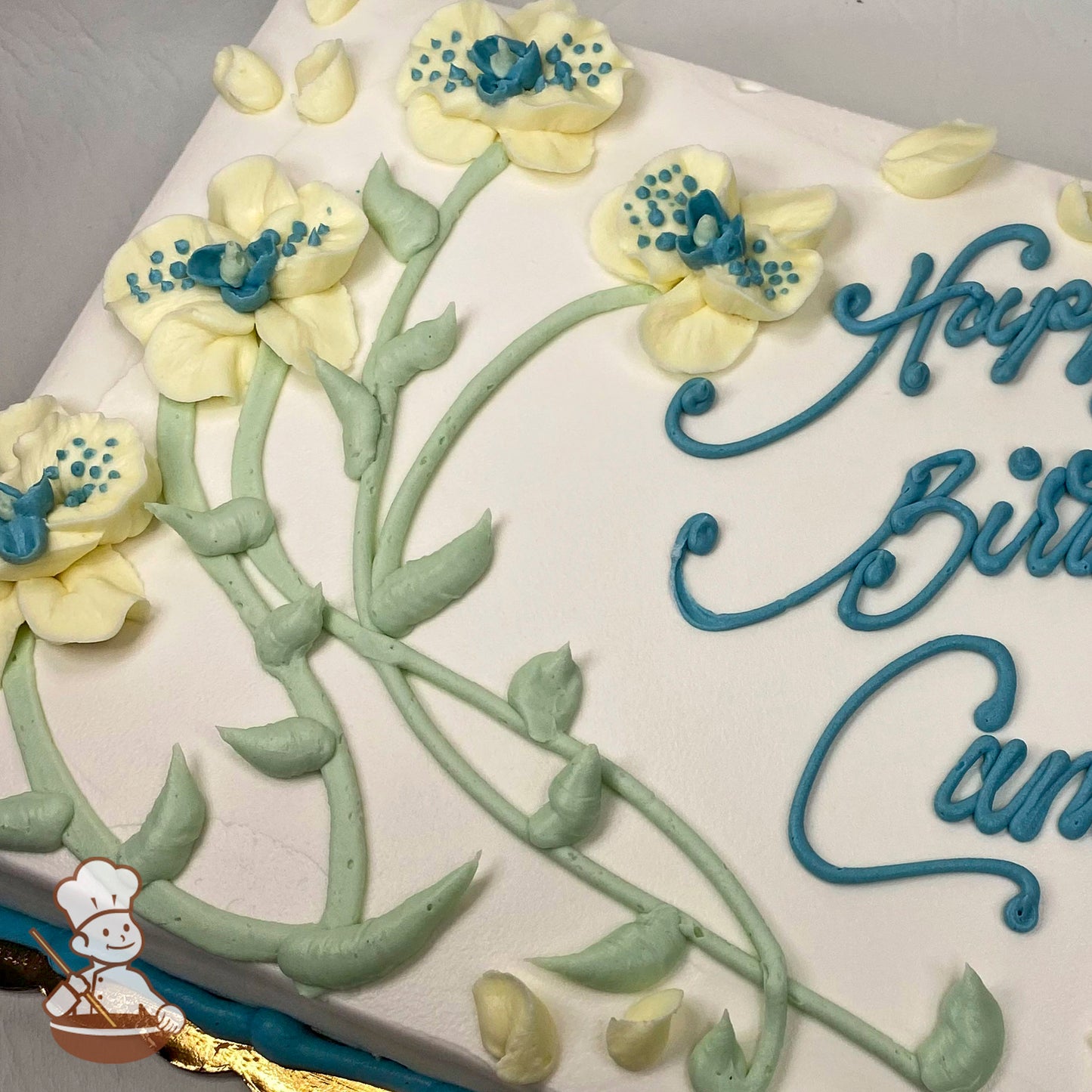 Sheet cake with white frosting, decorated with pale yellow flowers featuring blue centers, green stems, and leaves piped in buttercream.