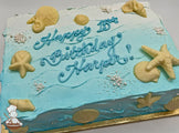 Sheet cake with blue ombré frosting, decorated with white chocolate seashells, starfish, sugar pearls, and wave-like frosting patterns.