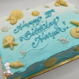 Sheet cake with blue ombré frosting, decorated with white chocolate seashells, starfish, sugar pearls, and wave-like frosting patterns.