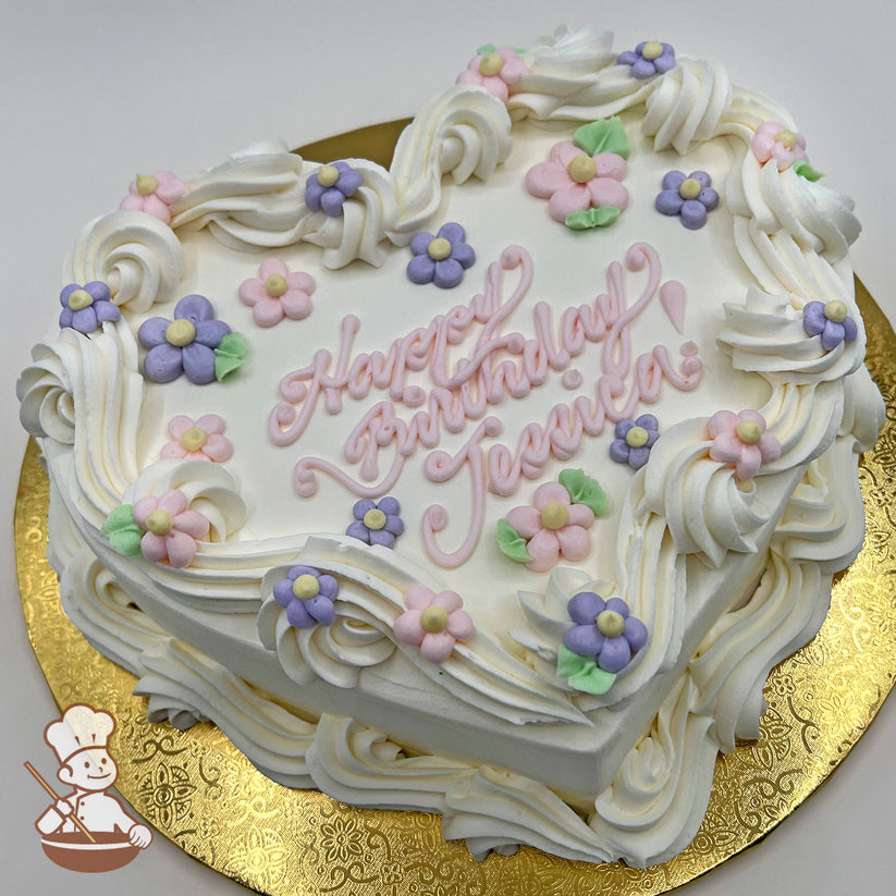 Heart-shaped cake with white buttercream swirls, pastel pink and purple daisy decorations, and green leaves on a gold board.