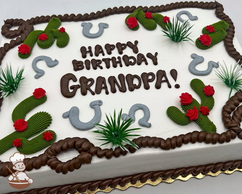 Sheet cake with white frosting, decorated with green buttercream cacti with red flowers, gray horseshoes, brown rope trim, and plastic green grass accents.
