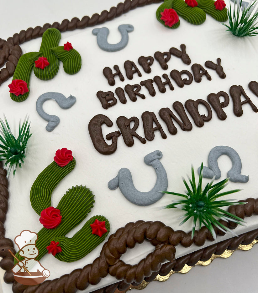 Sheet cake with white frosting, decorated with green buttercream cacti with red flowers, gray horseshoes, brown rope trim, and plastic green grass accents.