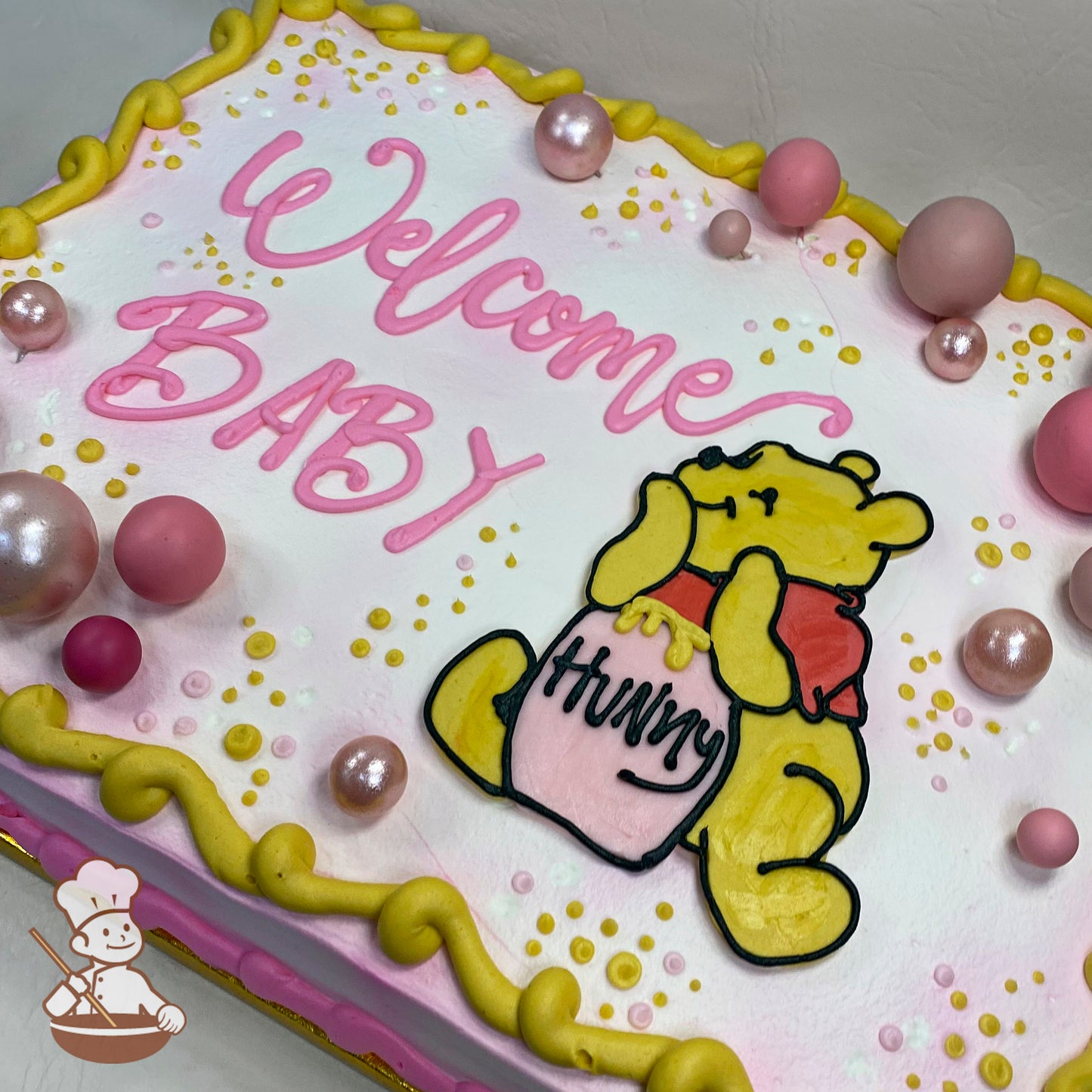 Sheet cake with pink and white frosting, yellow border, pink and rose gold bubble toppers, and a buttercream Winnie the Pooh holding a "Hunny" pot.