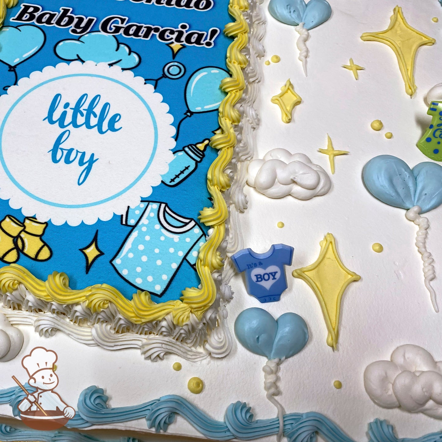 Sheet cake with white frosting, yellow and blue piped borders, blue heart-shaped balloon decorations, yellow star accents, and a plastic ring "It's a Boy" onesie decoration.