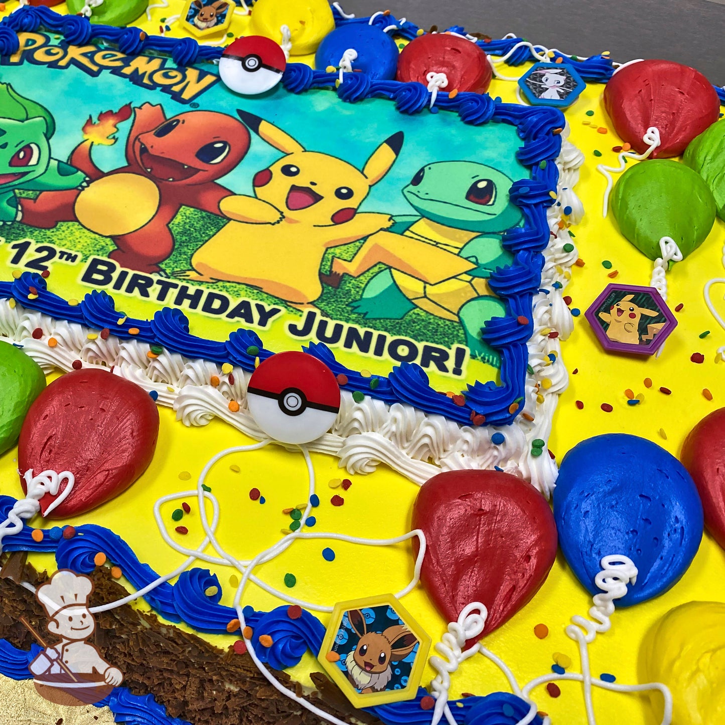 A yellow sheet cake with blue and white buttercream borders, decorated with red, blue, and green balloon designs, plastic Poké Ball toppers, and plastic Pokémon character decorations.