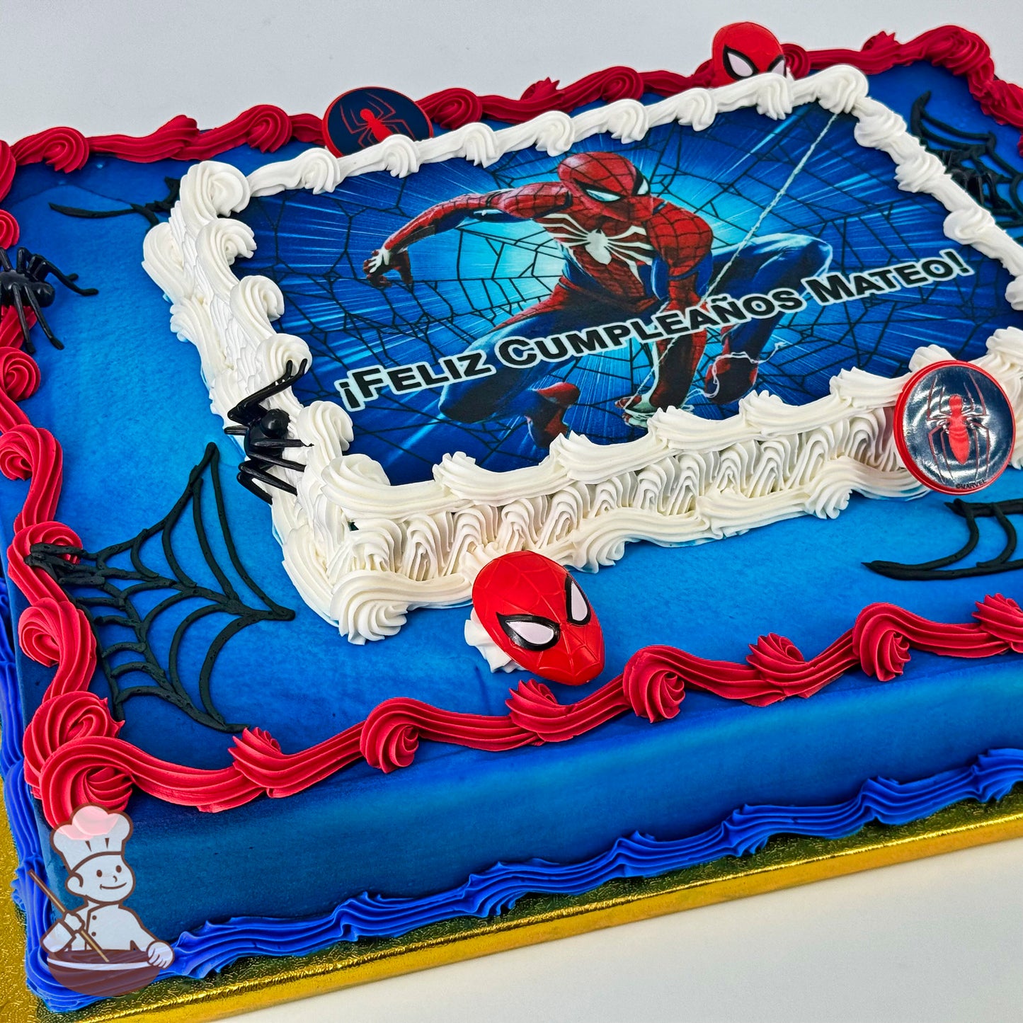 Sheet cake with blue base, red and blue piped borders, black spider web designs, plastic Spider-Man face and logo decorations, and white piped frosting around a central printed image.