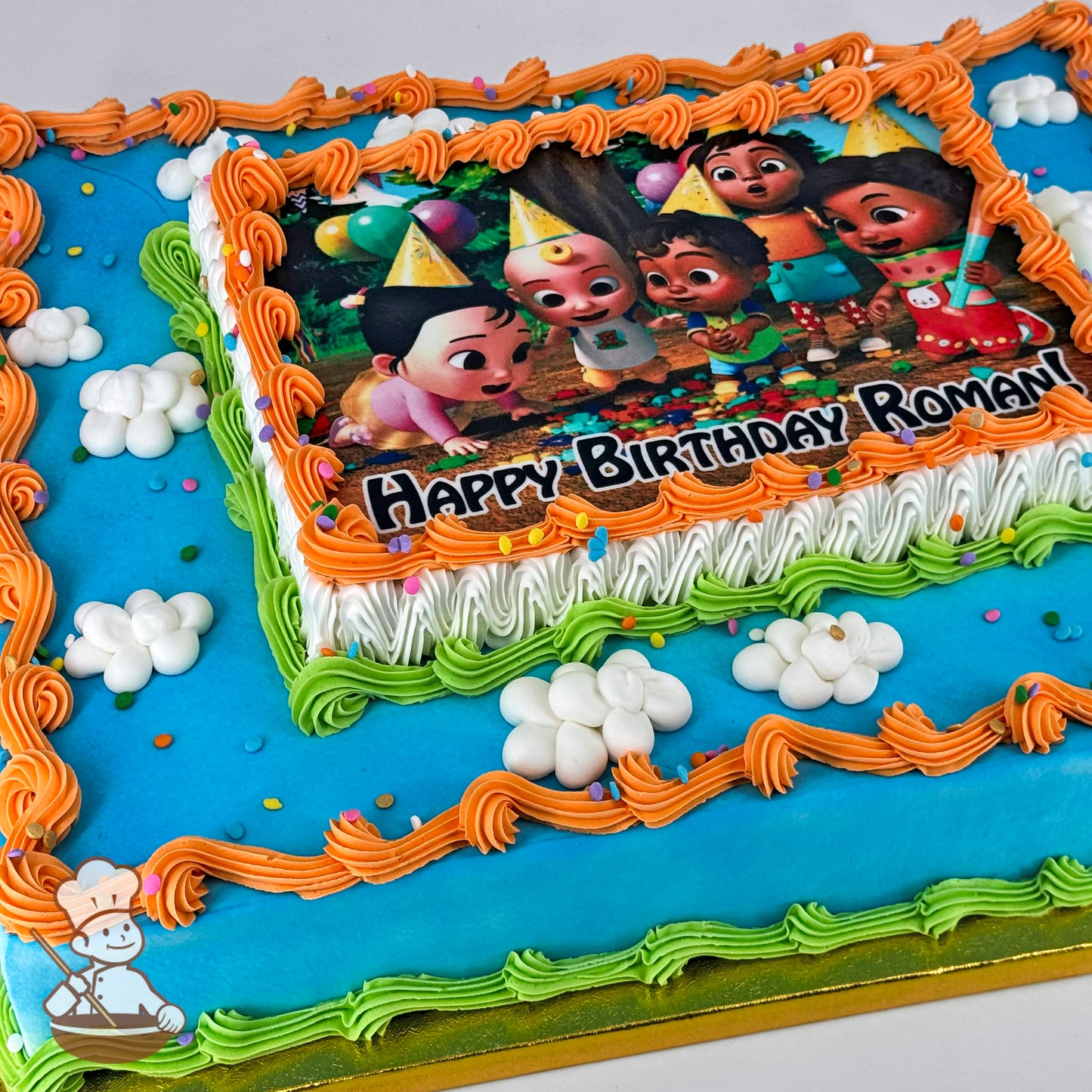 A rectangular sheet cake with blue frosting, orange and green piped borders, white buttercream clouds, and colorful sprinkles.