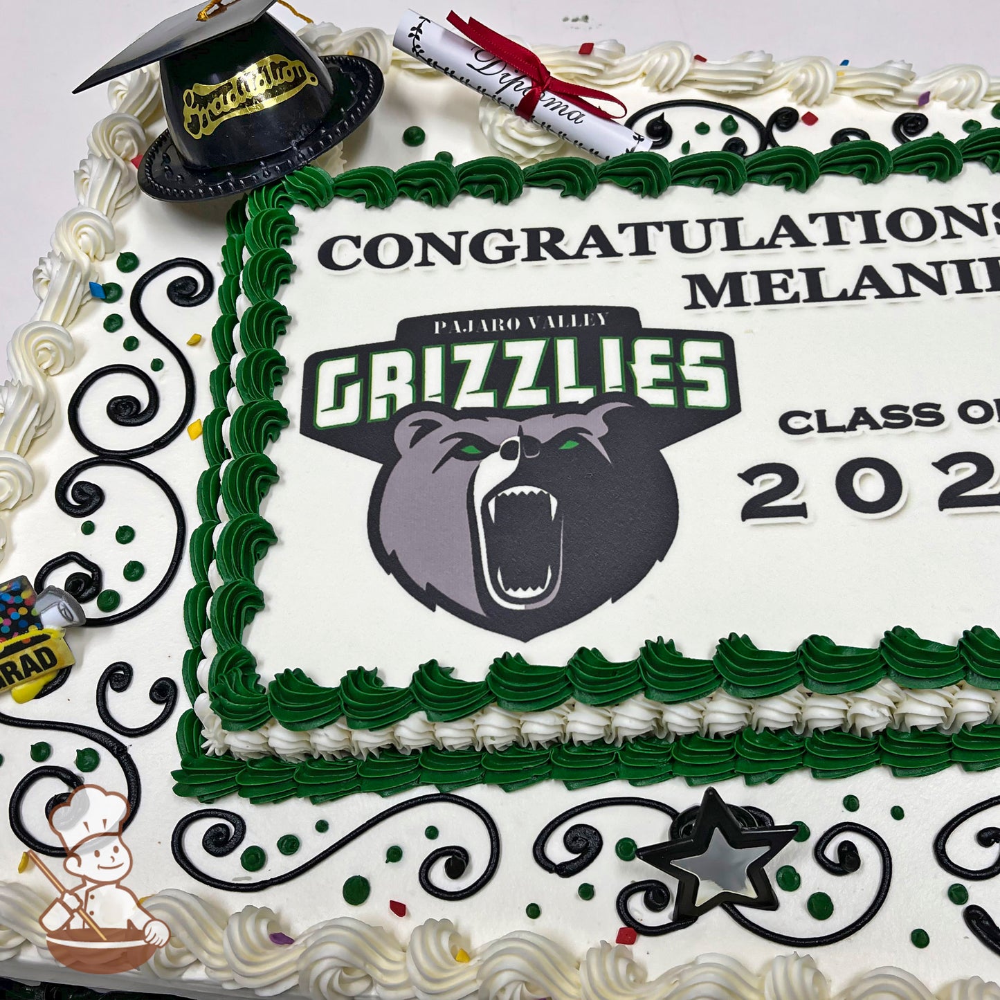 Sheet cake with green and white buttercream borders, black swirl designs, and plastic decorations of a graduation cap and diploma.
