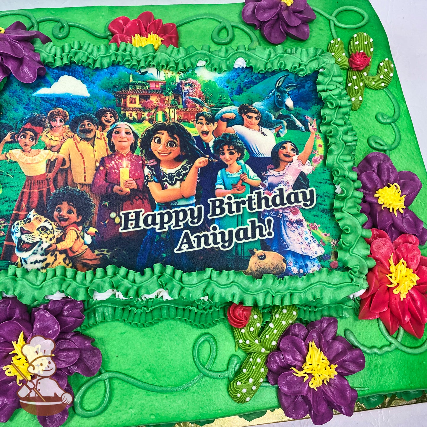 A green sheet cake with purple and hot pink buttercream flowers, yellow centers, green piped vines, and small cactus decorations surrounding a printed photo layer.