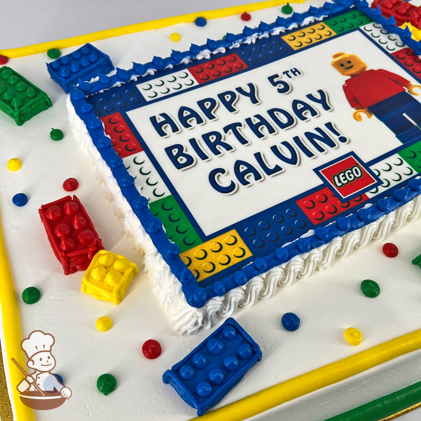 Sheet cake with a Lego-themed design, featuring colorful buttercream Lego brick decorations in red, blue, yellow, and green, and scattered dots.