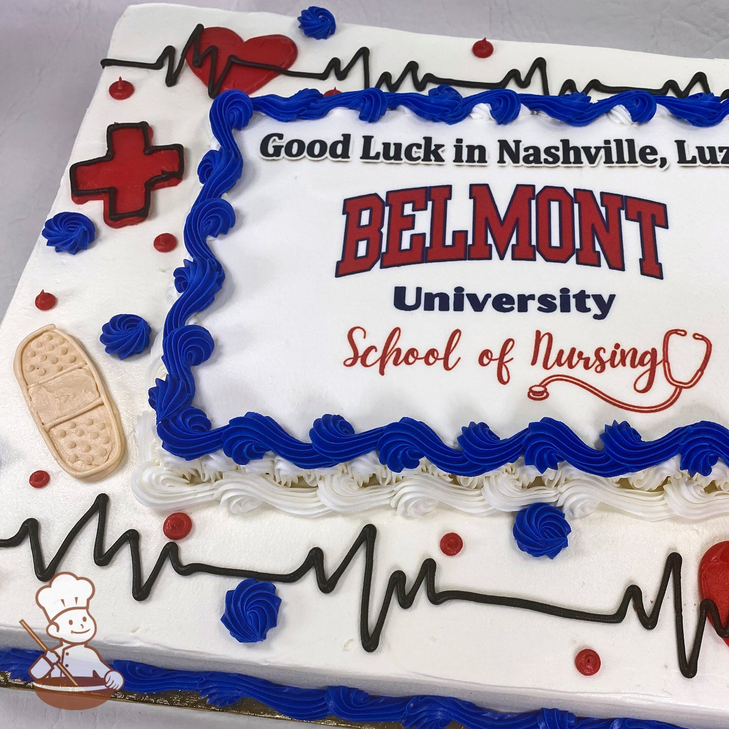 Sheet cake with white frosting, blue buttercream swirl trims, black piped heart rate lines, red heart and cross symbols, red dots, and a beige bandage decoration.