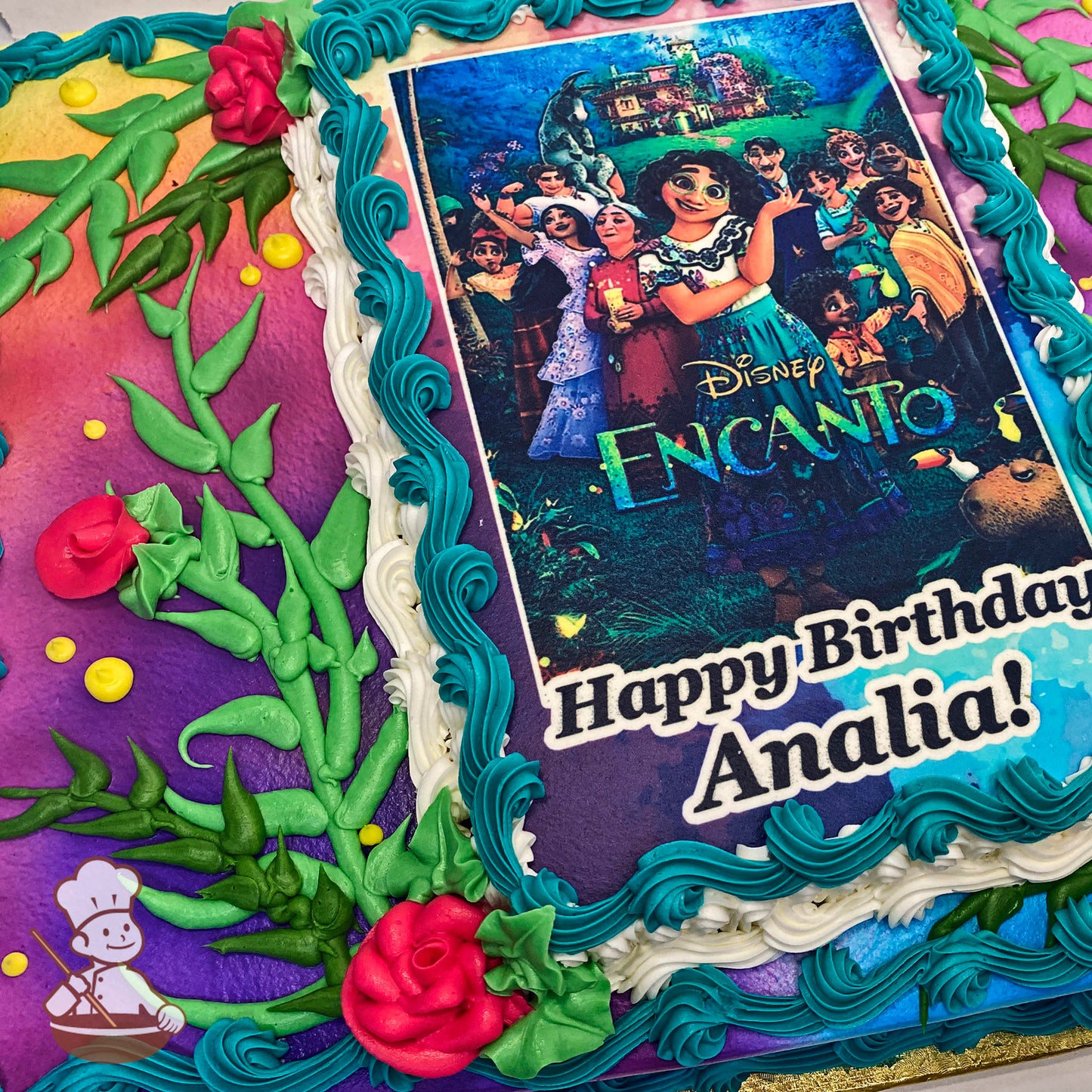 Sheet cake with colorful icing, featuring pink roses, green leaves, and vines. Blue and white borders surround a printed image.