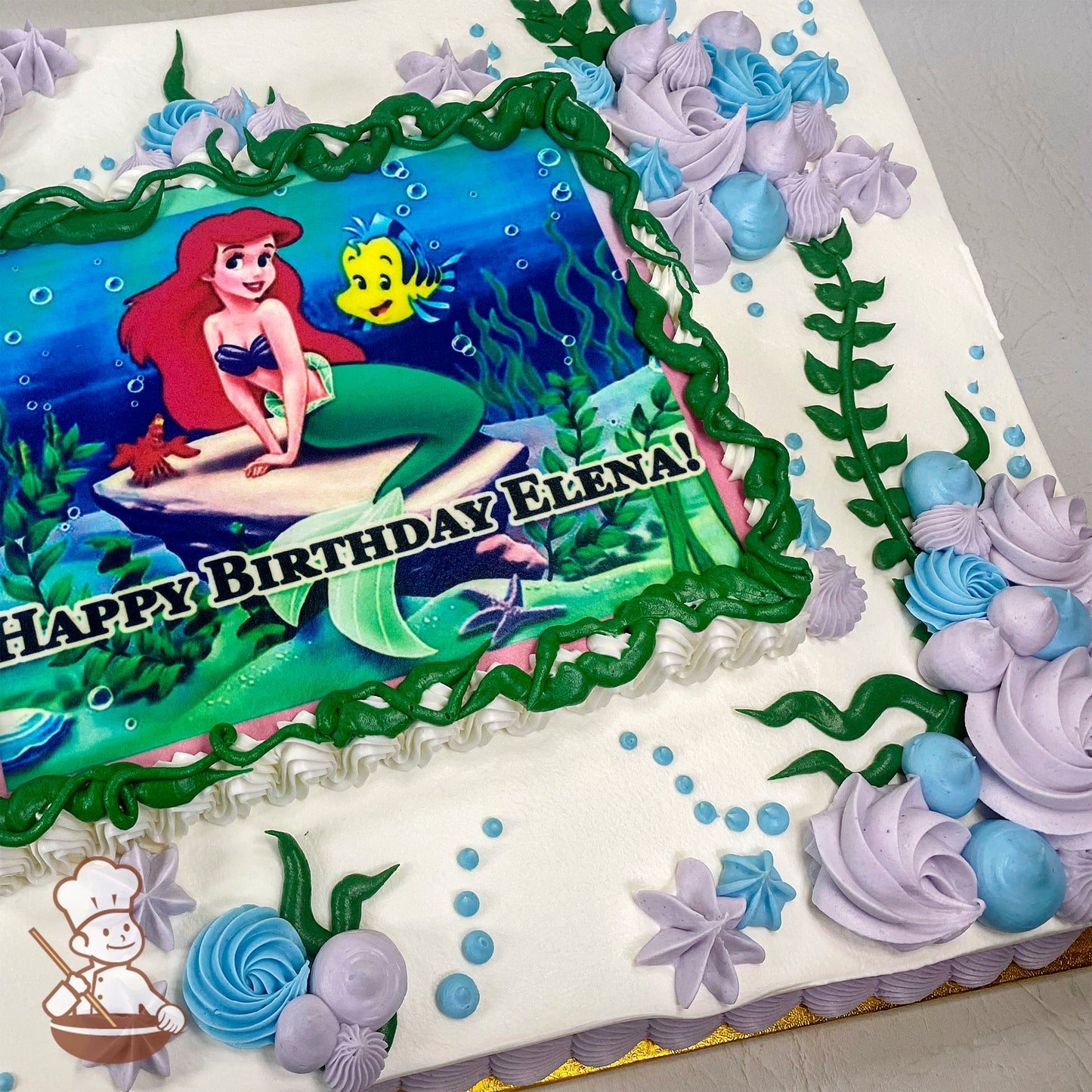 Sheet cake with a printed photo layer framed by green buttercream swirls, decorated with purple and blue buttercream swirls, star shapes, kelp, and bubbles.