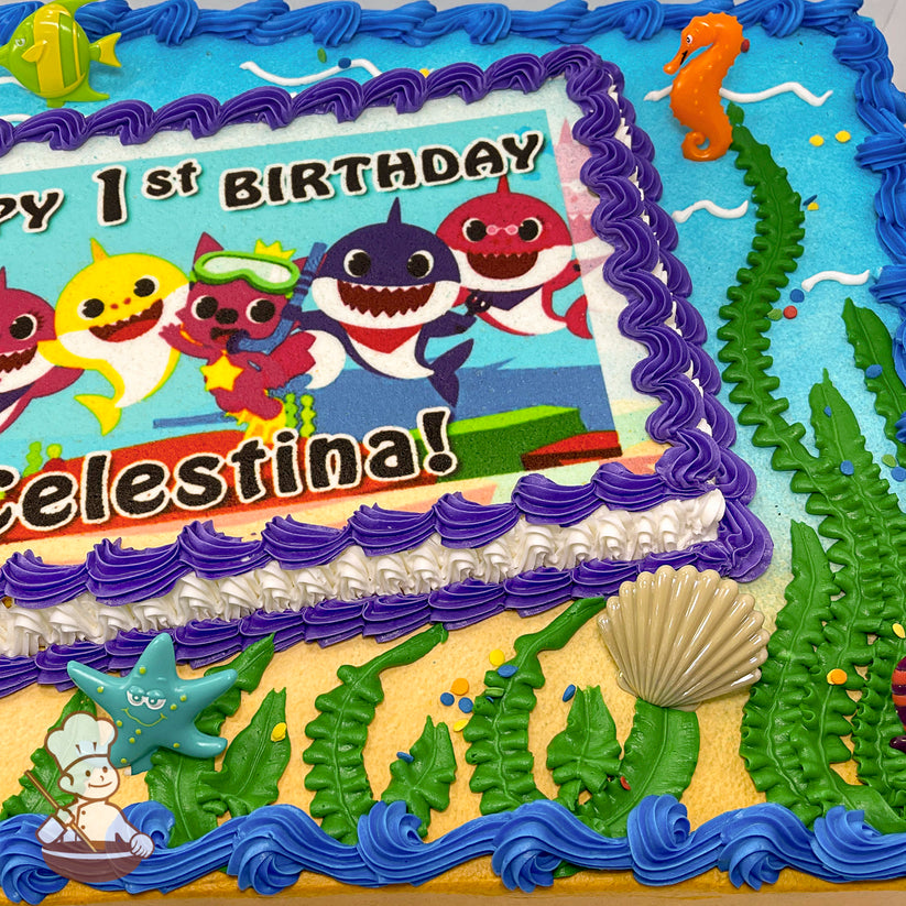 Sheet cake with a printed photo layer bordered by purple and white buttercream swirls. Decorations include green kelp, a plastic orange seahorse, a green fish, a blue starfish, a beige seashell, blue buttercream waves, and colorful sprinkles.