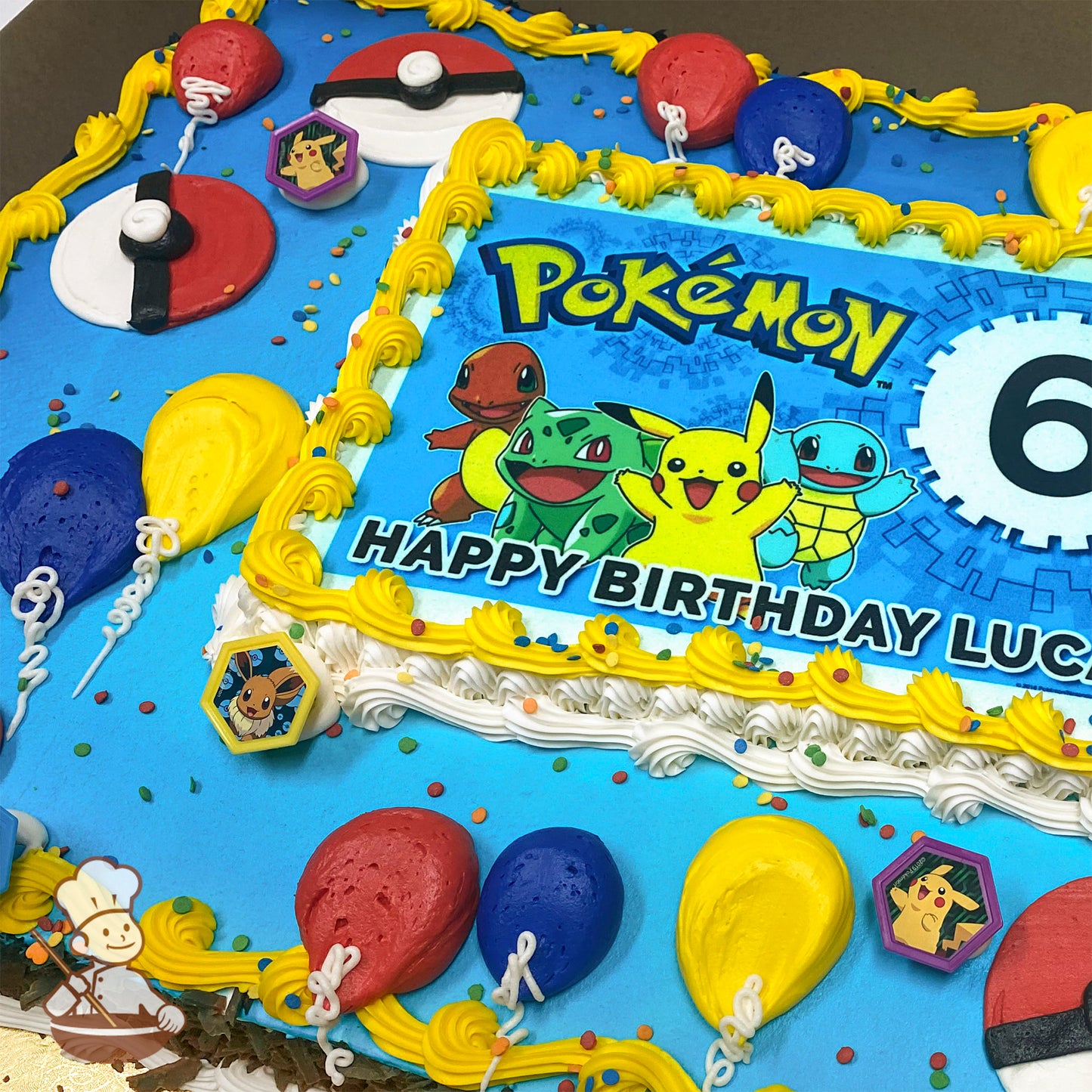 Sheet cake with blue frosting, yellow and white buttercream borders, balloon decorations in red, yellow, and blue, Poké Ball designs, and plastic Pokémon character rings featuring Pikachu and Eevee.