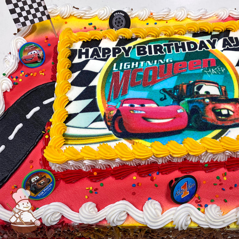Sheet cake with a printed "Cars" image, yellow and white buttercream borders, a black icing road, colorful sprinkles, and plastic character rings featuring Lightning McQueen, Mater, and a "Rust-eze" logo.