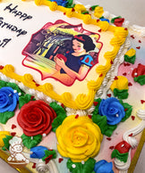 Sheet cake with buttercream roses in red, yellow, and blue, green leaves, white and yellow swirl borders, and red heart sprinkles around the edges.