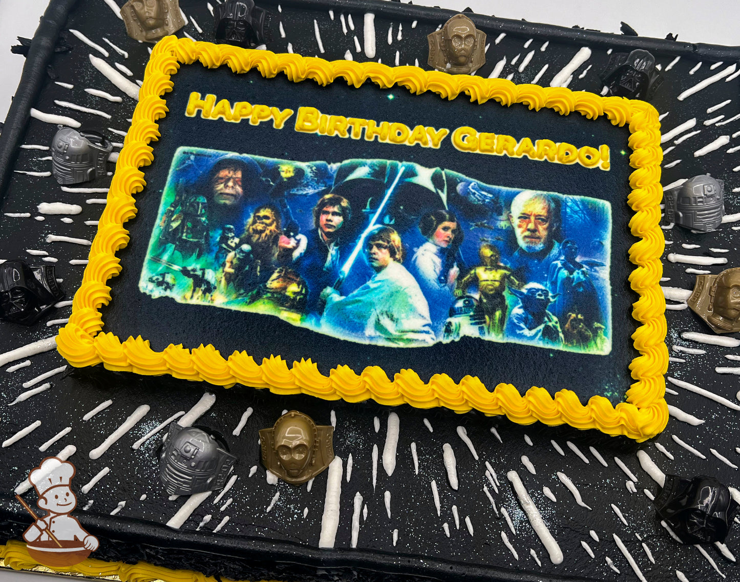 Sheet cake with a Star Wars theme, featuring a yellow buttercream border around a printed image, black frosting with white streaks resembling hyperspace, and plastic Star Wars character rings as decorations.