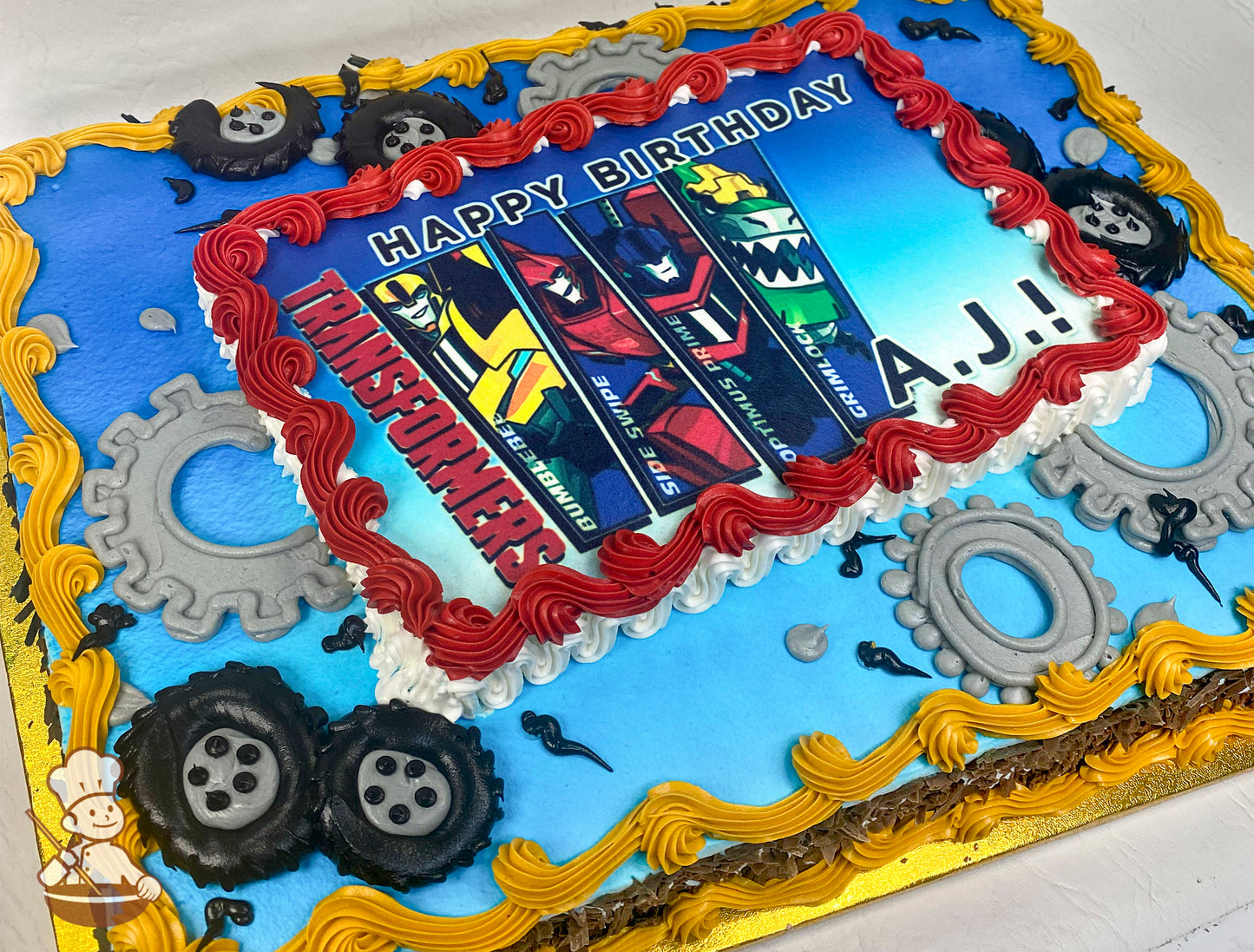 Sheet cake with a blue base, red and yellow piped borders, gray gear decorations, and black piped tires around the edges.