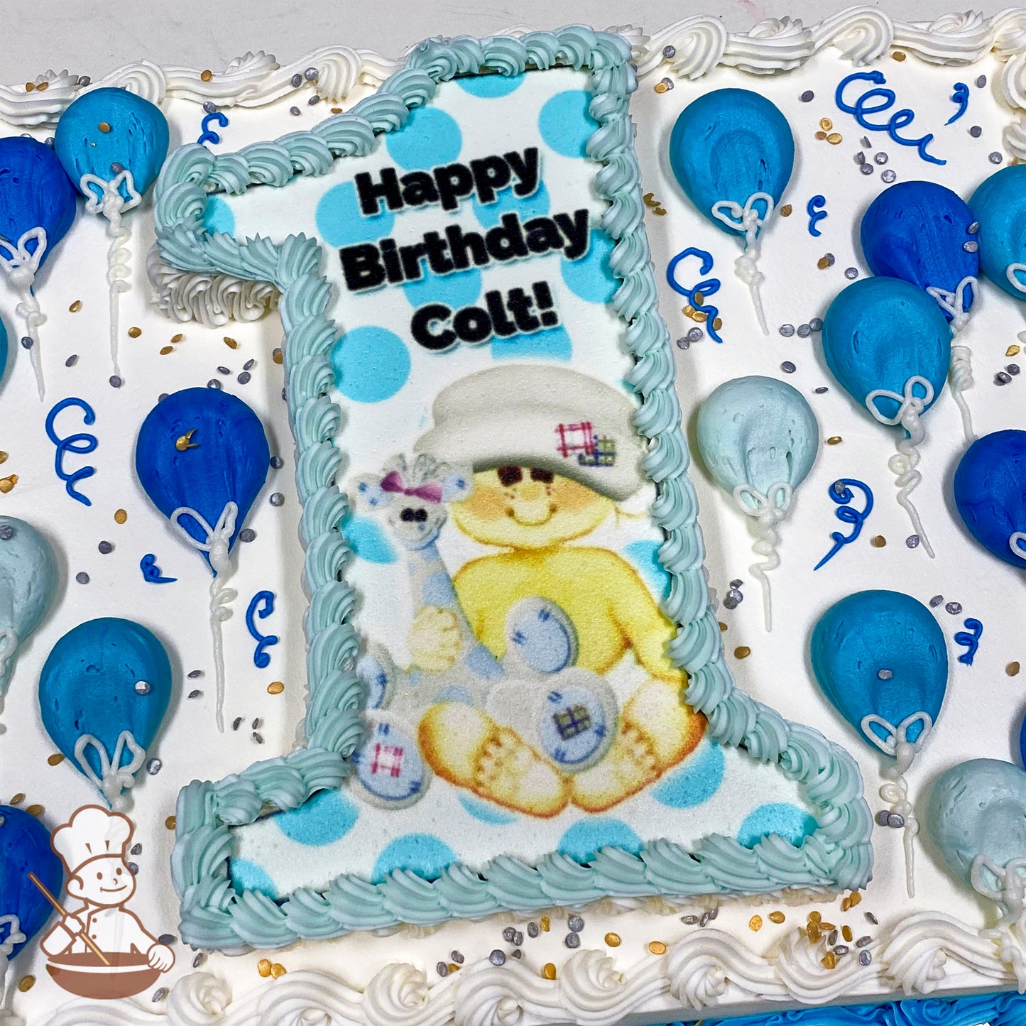 Sheet cake with buttercream decorations, featuring a large number "1" cutout with a baby boy holding a giraffe design. Blue balloons, confetti sprinkles, and swirls decorate the cake.