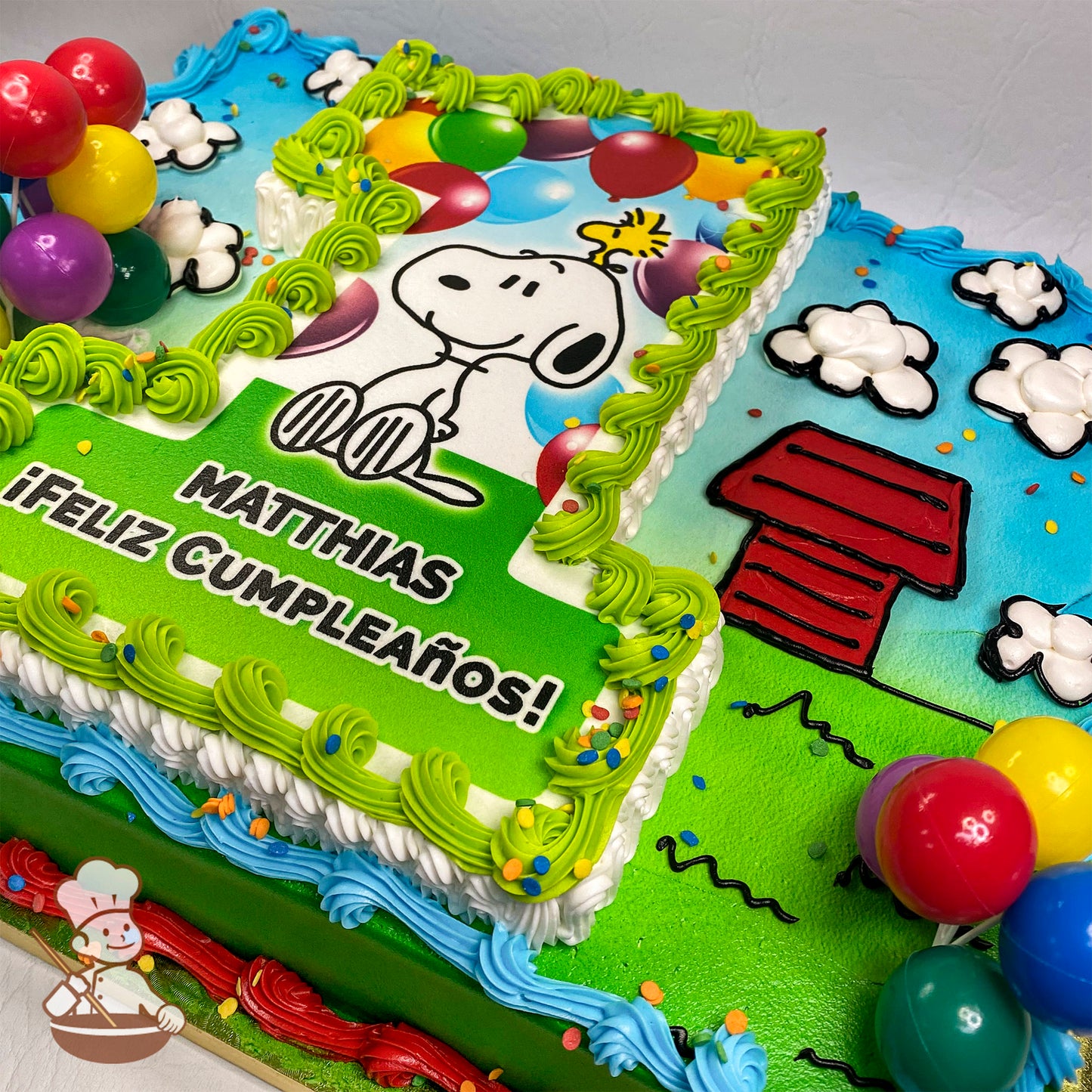 Sheet cake with a printed Snoopy design bordered by green buttercream swirls, red doghouse, puffy buttercream clouds, colorful balloons, and confetti accents.
