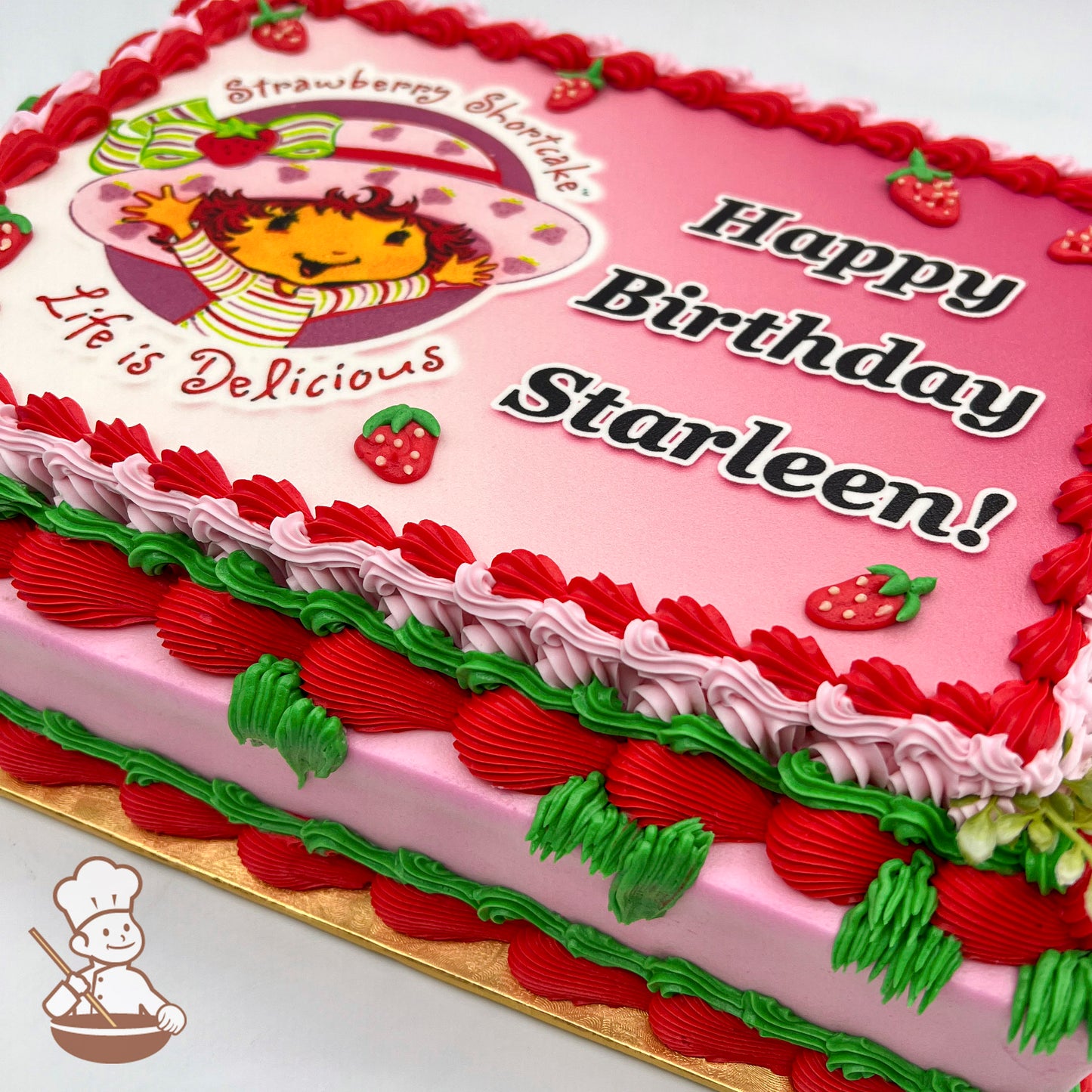 Sheet cake with pink frosting, red and green buttercream piped strawberries, and a red scalloped border.