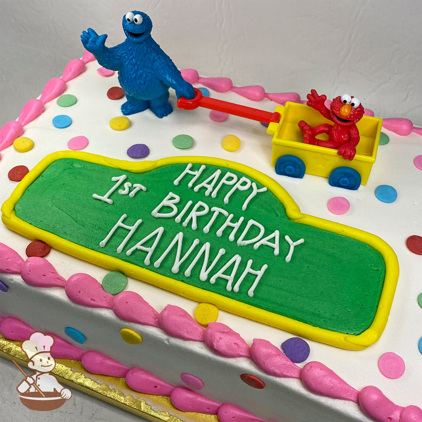Sheet cake with pink buttercream borders, colorful polka dots, a green buttercream plaque, and plastic Cookie Monster pulling Elmo in a yellow wagon.