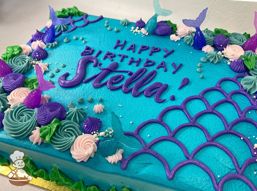Blue sheet cake with plastic mermaid tail toppers, buttercream rosettes in pink, teal, and purple, green leaves, white pearl sprinkles, and purple scallop designs.