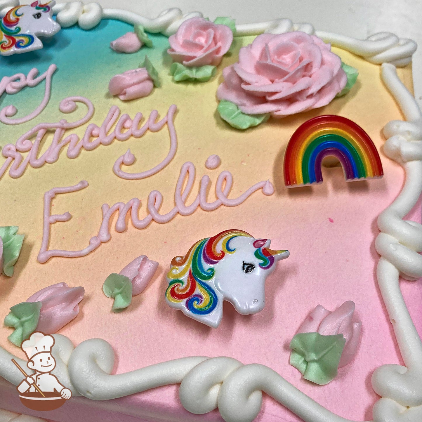 Sheet cake with a pastel rainbow gradient background, pink buttercream roses, green leaves, plastic unicorn and rainbow decorations, and white frosting borders.