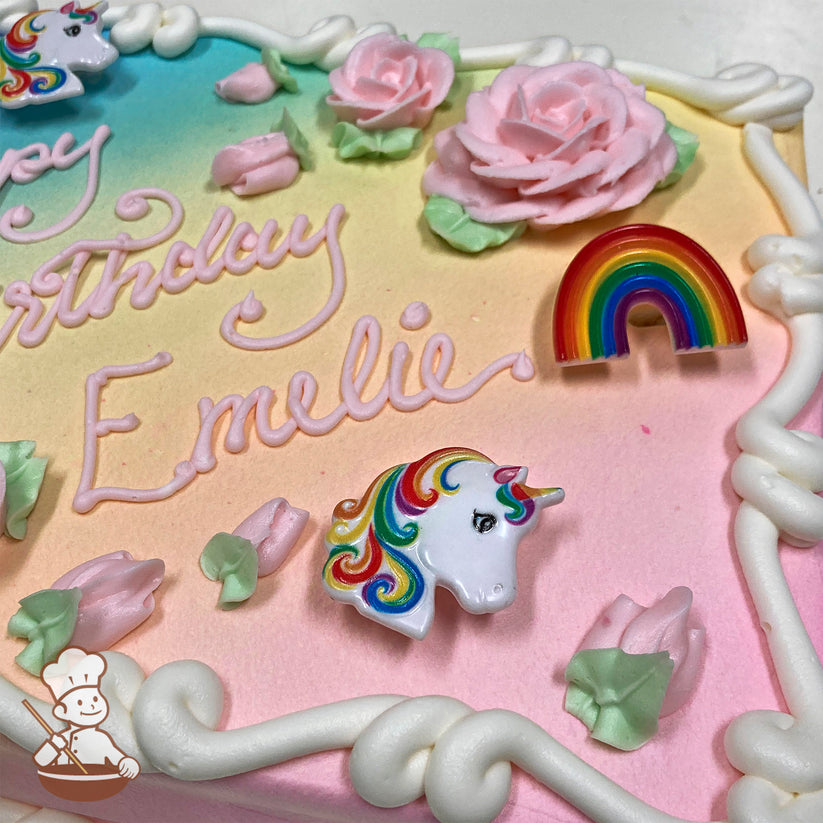 Sheet cake with a pastel rainbow gradient background, pink buttercream roses, green leaves, plastic unicorn and rainbow decorations, and white frosting borders.