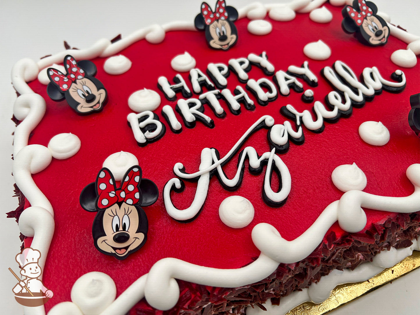 Red sheet cake with white buttercream dollops, swirl trims, and Minnie Mouse plastic rings featuring her face and red polka-dot bow as decorations.