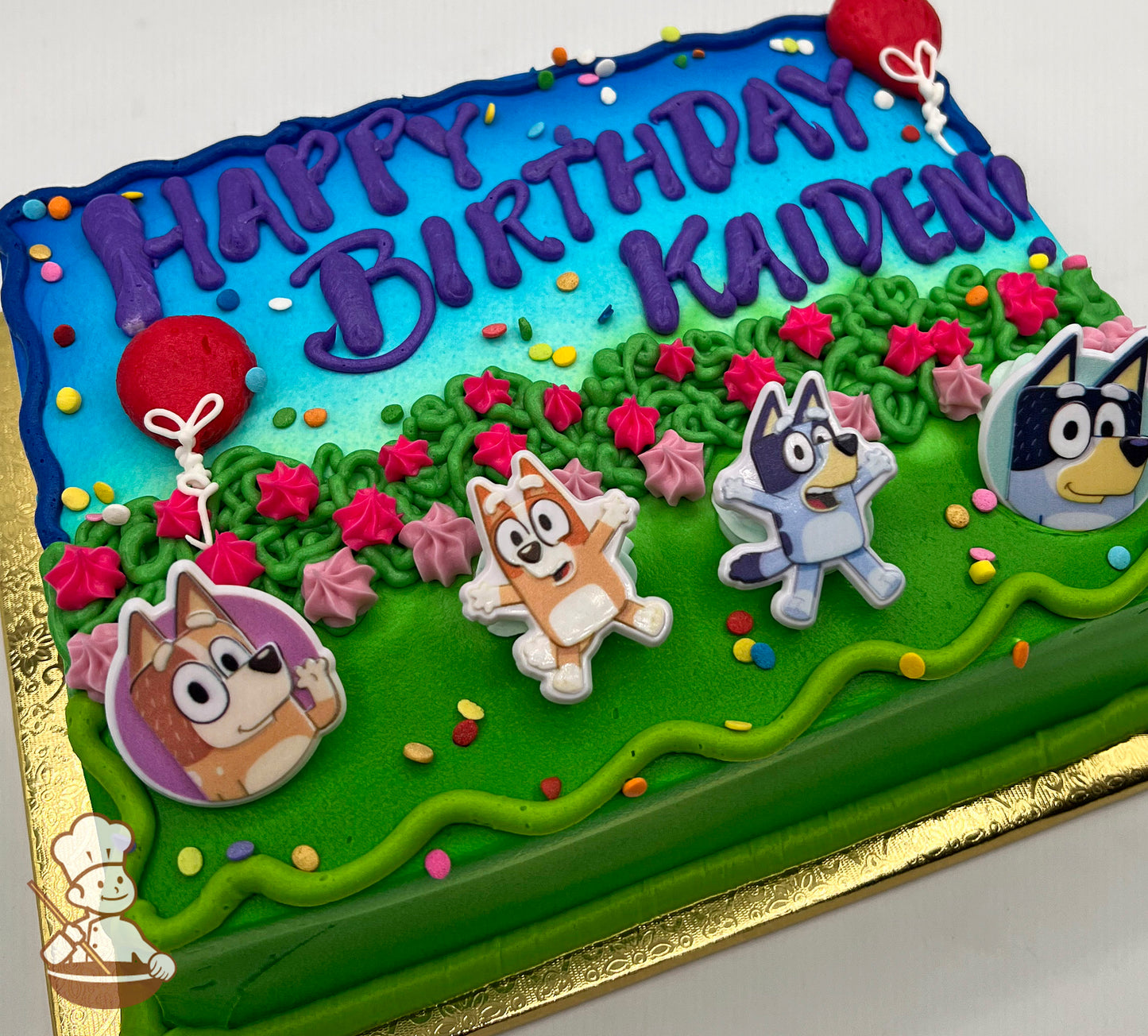Sheet cake with a blue and green design, featuring plastic Bluey character toppers, pink and green floral accents, and colorful sprinkles.