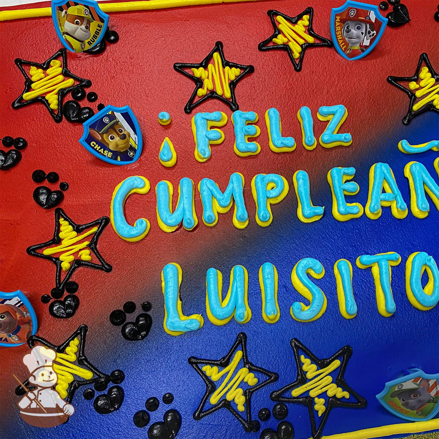 Sheet cake with red and blue frosting, yellow and black star designs, black paw prints, and plastic Paw Patrol character badges featuring Rubble, Chase, and Marshall.