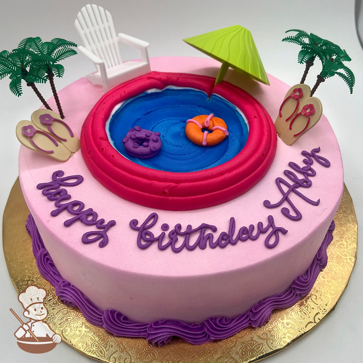 Pink round cake with a buttercream pool design, plastic palm trees, a green umbrella, a white chair, sandal decorations, and float rings.