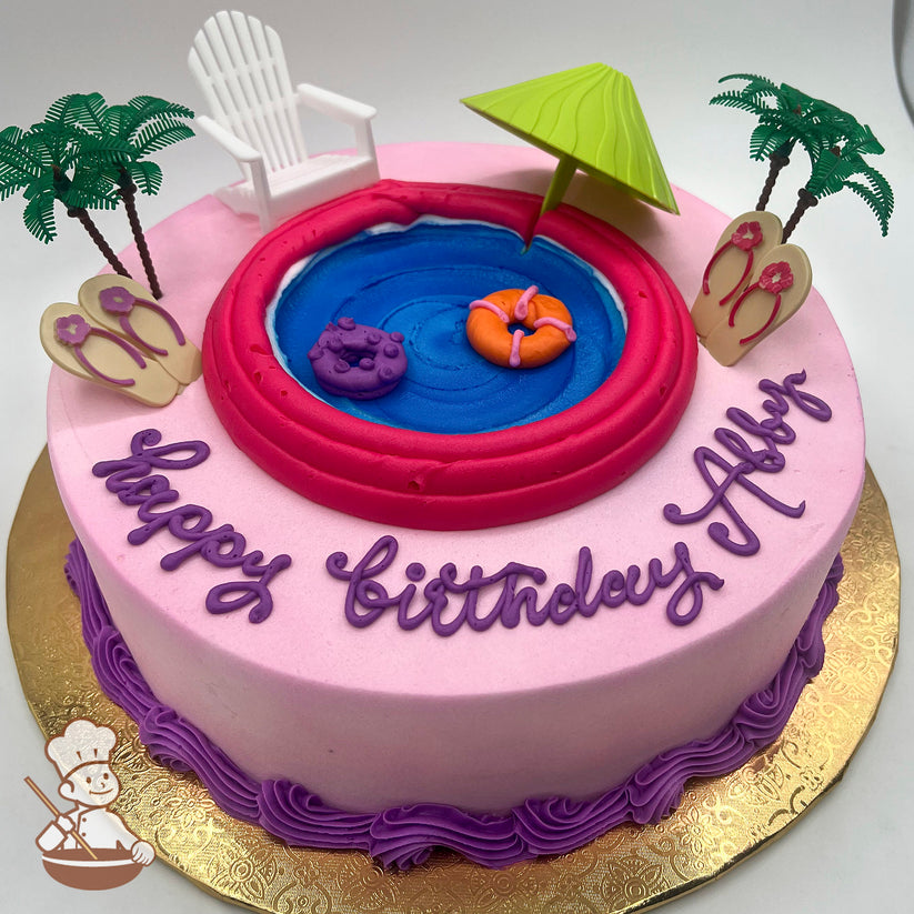 Pink round cake with a buttercream pool design, plastic palm trees, a green umbrella, a white chair, sandal decorations, and float rings.